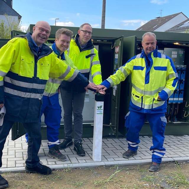 Vier Männer in blau-gelber Arbeitskleidung stehen vor einem grünen Kasten mit geöffneten Türen: der digitalen Strom-Ortsnetzstation in Sankt Augustin-Menden.