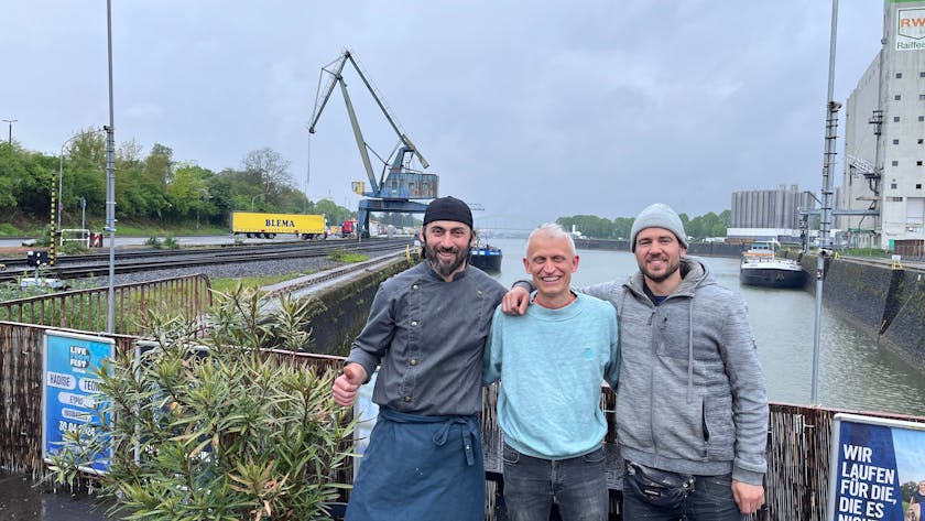 Erkan Karakas, Inhaber der Erkantine, Philip Minettos vom Verein Rosarot und Unterstützer Mario Sroka freuen sich auf das „Vrühlingsfest“ am Niehler Hafen.