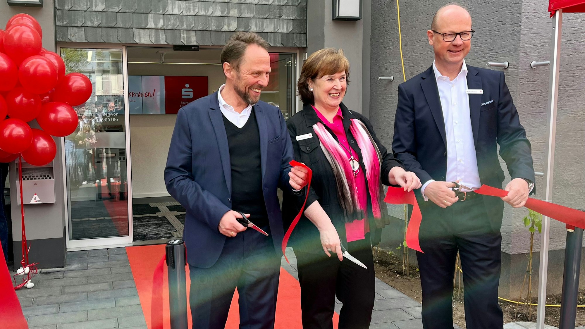 OB Uwe Richrath, Filialleiterin Heike Schulz und Sparkassenvorstand Markus Grawe schneiden vor der umgebauten Filiale in Lützenkirchen ein rotes Band durch.