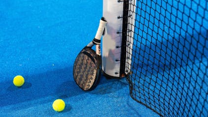 Das Bild zeigt einen Padel-Schläger, der an dem Pfosten eines Tennisnetzes lehnt. Auf dem blauen Court liegen zwei gelbe Tennisbälle.