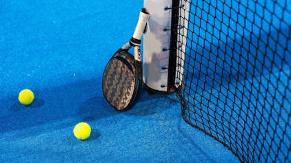Das Bild zeigt einen Padel-Schläger, der an dem Pfosten eines Tennisnetzes lehnt. Auf dem blauen Court liegen zwei gelbe Tennisbälle.