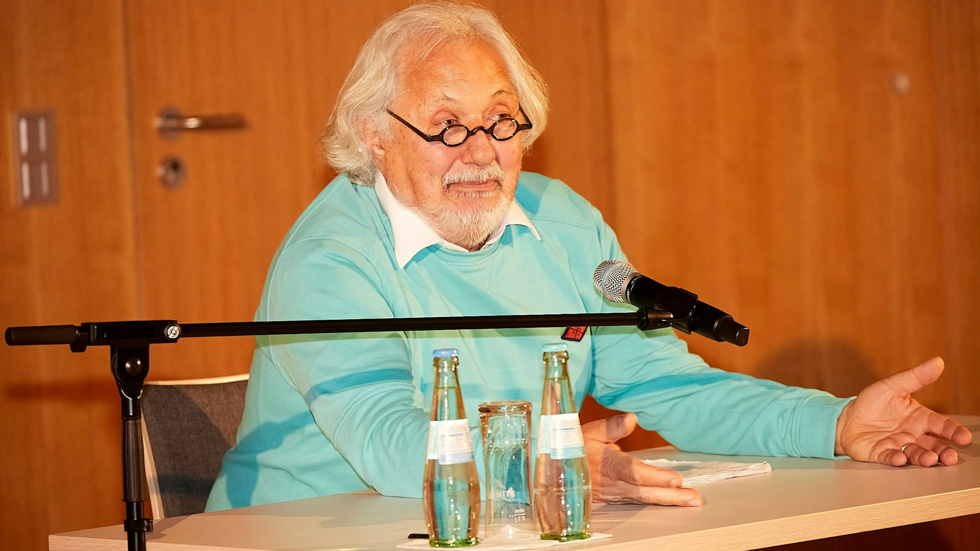 Konrad Beikircher trägt einen türkisfarbenen Pullover, er sitzt an einem Tisch und spricht in ein Mikrofon.