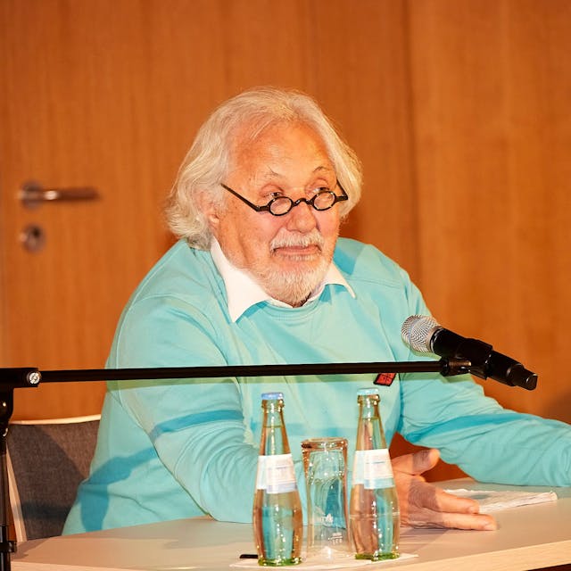 Konrad Beikircher trägt einen türkisfarbenen Pullover, er sitzt an einem Tisch und spricht in ein Mikrofon.