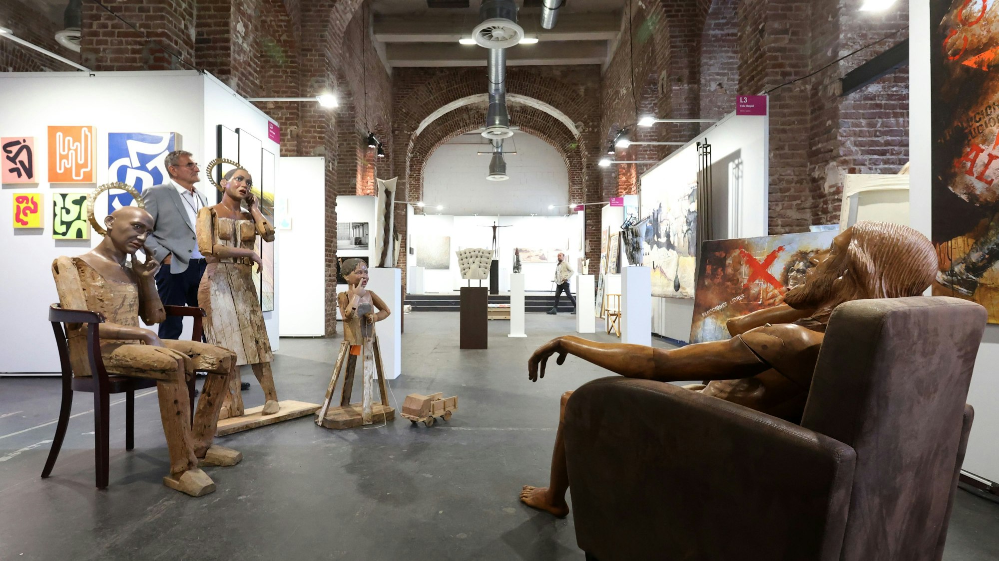In einer Halle sind lebensgroße Holz-Menschen ausgestellt, an den Wänden hängen Kunstwerke