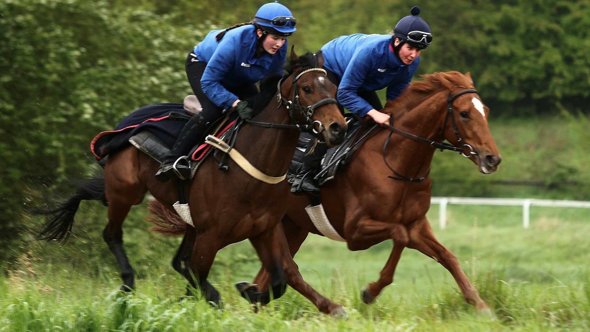 Zwei Jockeys sind auf Pferden in schnellen Tempo unterwegs. Im Hintergrund sieht man die Umrandungen einer Rennstrecke.