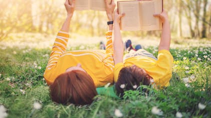 Mutter und Sohn liegen auf einer Gänseblümchen-Wiese im Frühling und lesen ein Buch. Symbolbild
