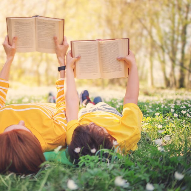 Mutter und Sohn liegen auf einer Gänseblümchen-Wiese im Frühling und lesen ein Buch. Symbolbild