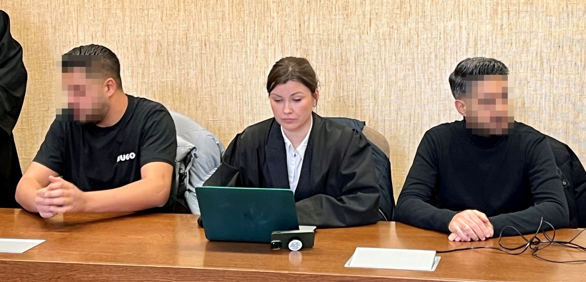 Zwei Männer sitzen auf der Anklagebank, zwischen ihnen eine Anwältin, die auf ihr Laptop guckt.
