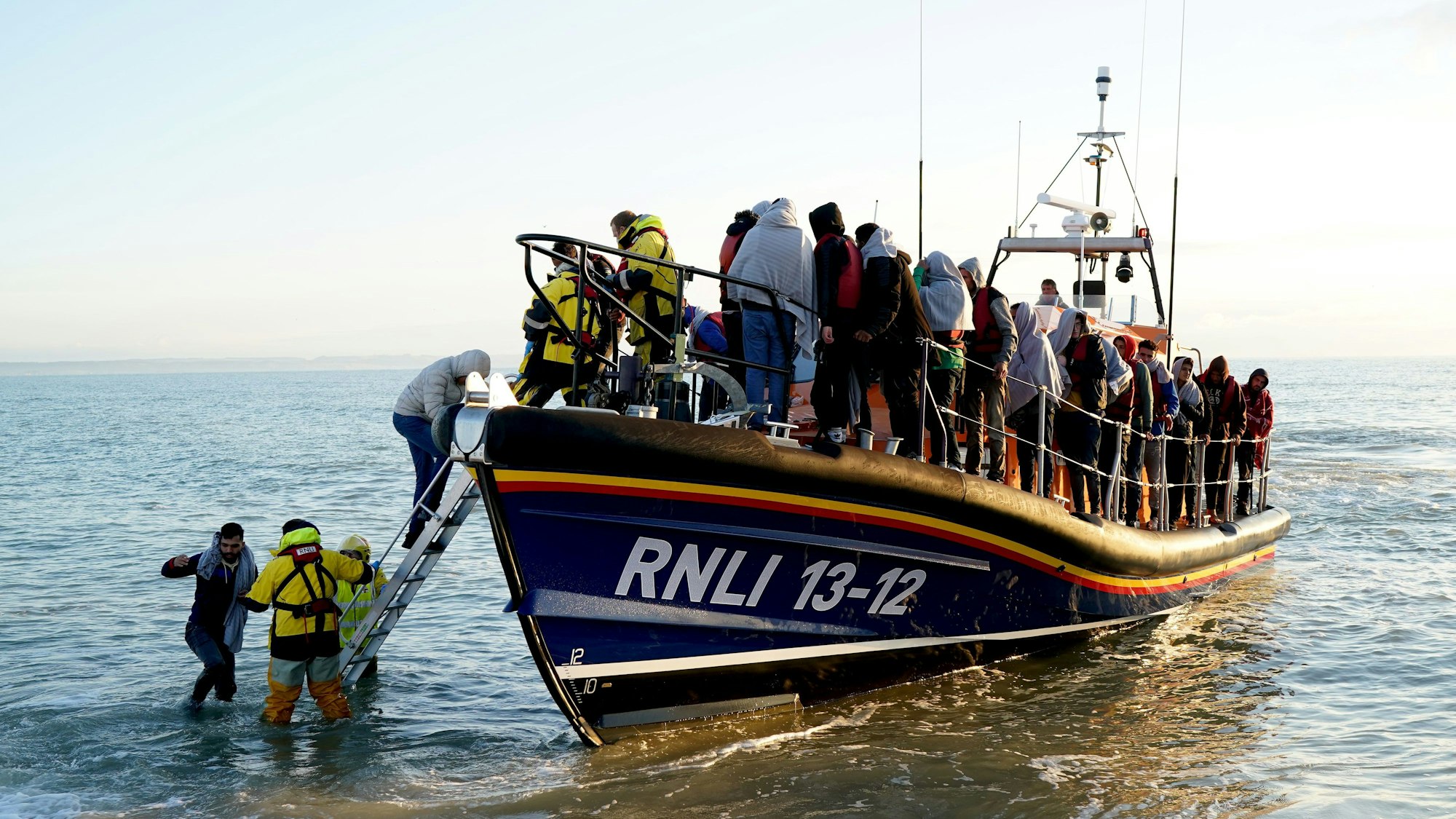 Eine Gruppe von Menschen auf einem kleinen Boot, bei denen es sich vermutlich um Migranten handelt, wird von der Royal National Lifeboat Institution (RNLI) nach einem Zwischenfall im Ärmelkanal nach Dungeness in der Grafschaft Kent gebracht. Großbritannien will illegal eingereiste Migranten künftig nach Ruanda abschieben