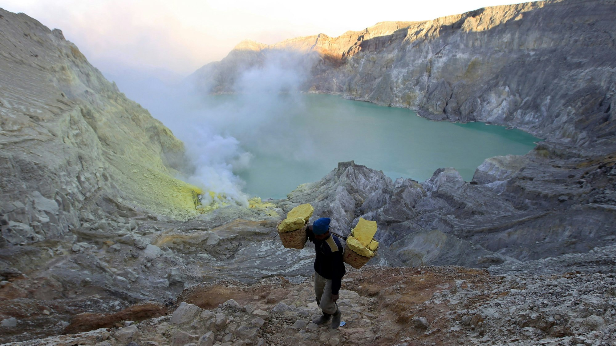 Ein Bergarbeiter trägt Körbe mit Schwefel. Im Hintergrund ist der Vulkankrater zu sehen.