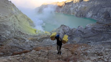 Ein Bergarbeiter trägt Körbe mit Schwefel. Im Hintergrund ist der Vulkankrater zu sehen.