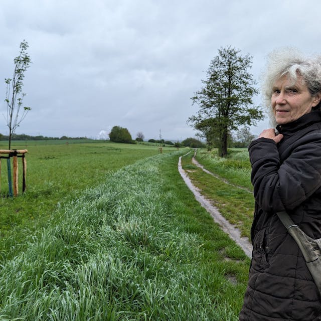 Die Ratsfrau Inge Cürten-Noack von den Grünen steht auf einem Feldweg vor einem Ackerstreifen, auf dem junge Bäume gepflanzt worden sind.