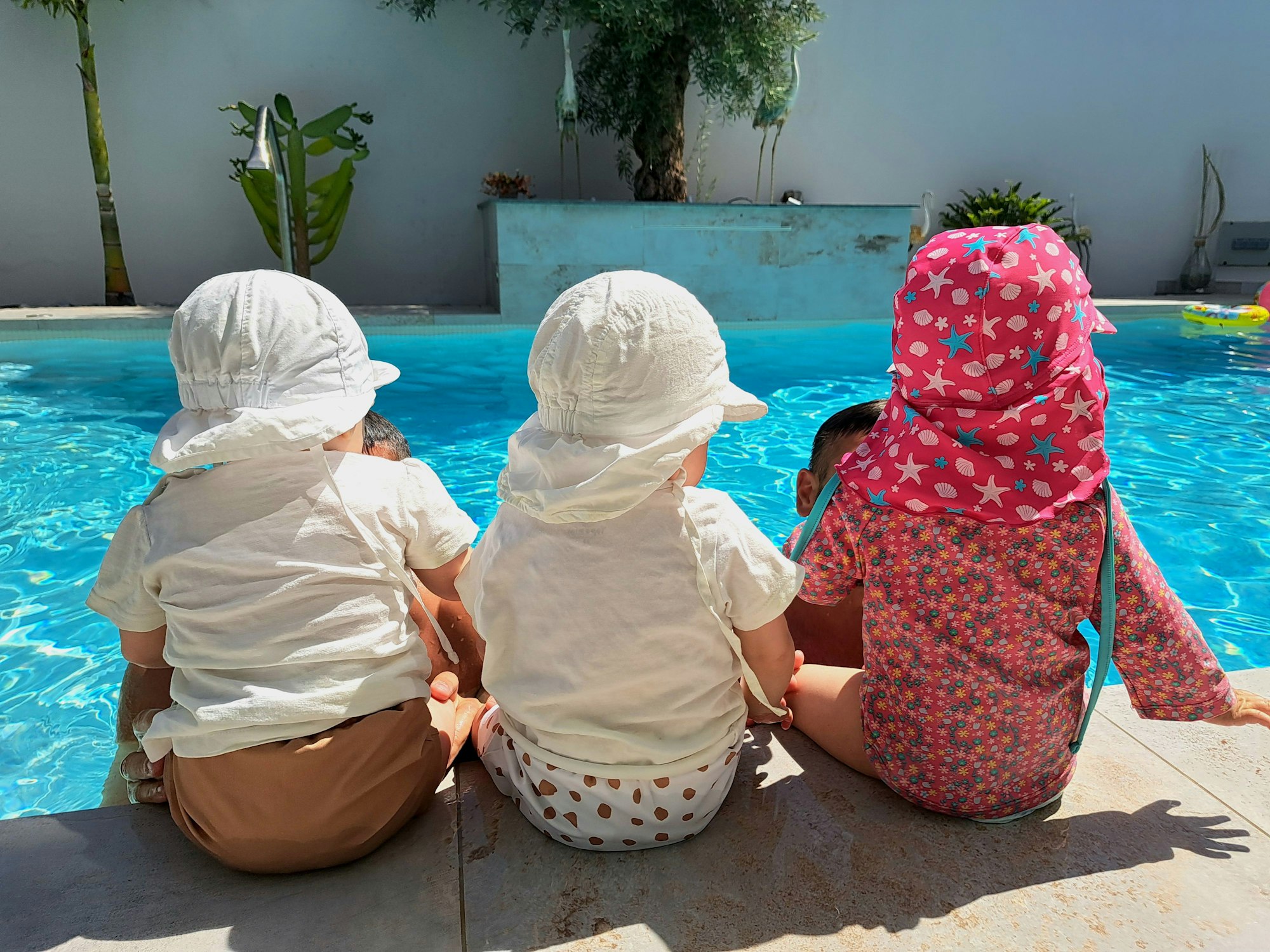 Drei Kleinkinder sitzen mit dem Rücken zum Betrachter an einem Pool nebeneinander.