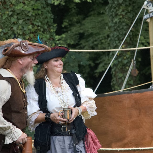 Ein Mann und eine Frau, selbst wie Piraten kostümiert, verfolgen vor einem Holzboot das Geschehen.