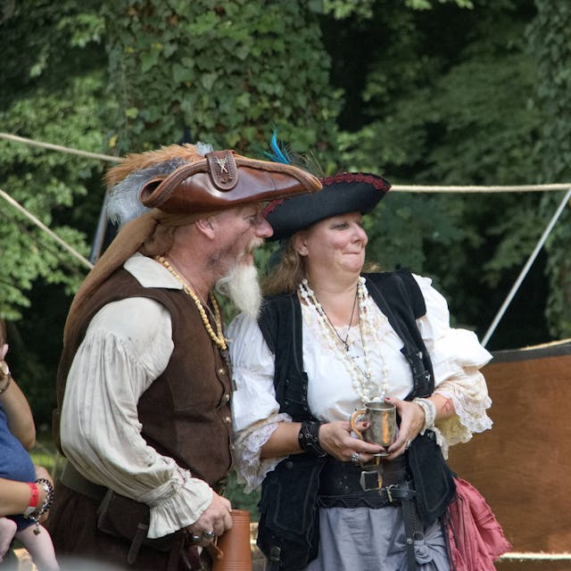 Ein Mann und eine Frau, selbst wie Piraten kostümiert, verfolgen vor einem Holzboot das Geschehen.
