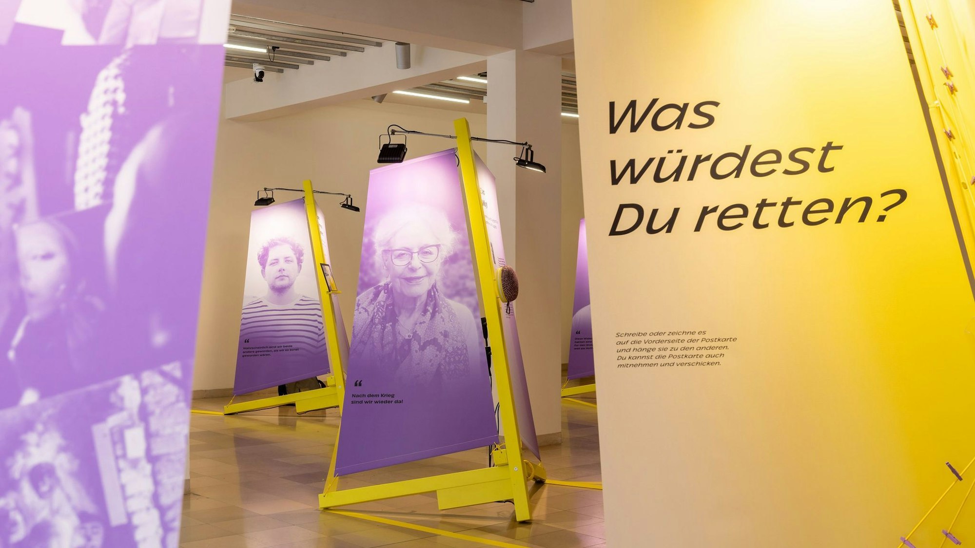 Violette Segel hängen an gelben Masten und zeigen die Kulturretter. Man sieht im Hintergrund etwa die Holocaust-Überlebende Eva Weyl. Im Vordergrund steht ein Segel mit der Aufschrift: „Was würdest du retten?“.