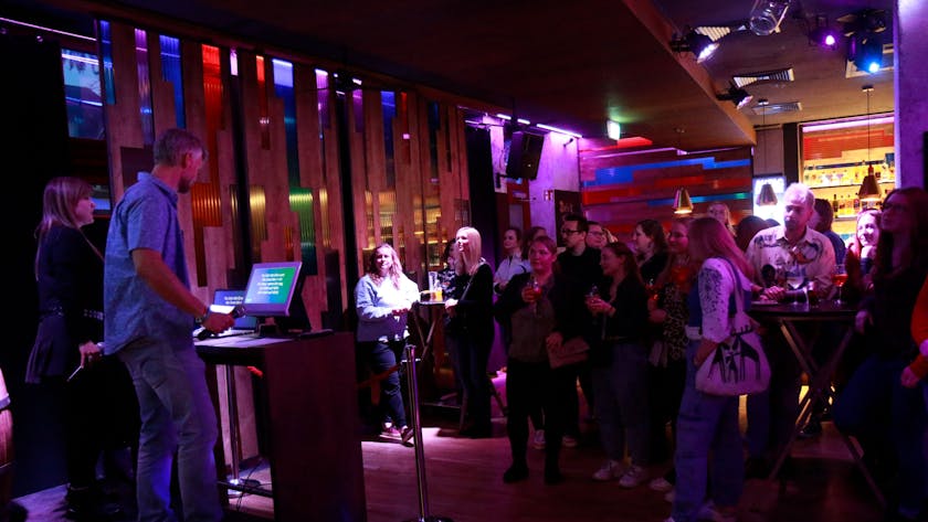 Eine Menschengruppe steht vor einem Pult in einem spärlich beleuchteten Raum und singt.