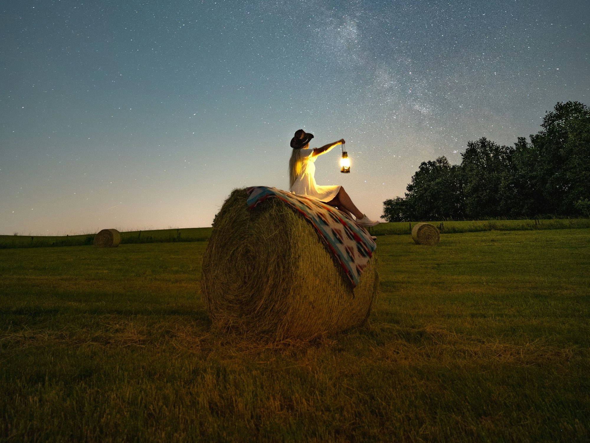 Eine Frau sitzt nachts auf einem Heuballen und hält eine leuchtende Laterne in die Höhe. Am Himmel sind zahlreiche Sterne zu erkennen.