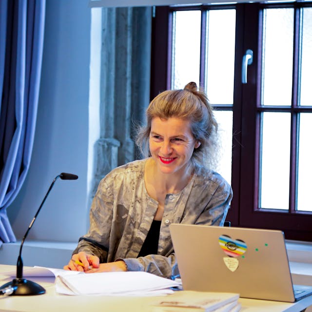 Anne Cotten sitzt an einem Schreibtisch, vor sich ein Stapel Papiere, ein Laptop und einem Mikrofon. Sie hat ihre Haare hinten zu einem Topf zusammengebunden.