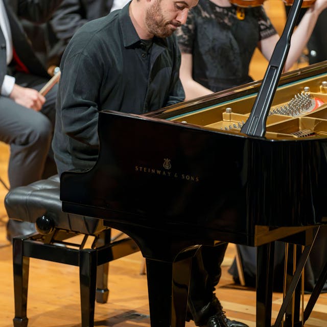 Auf dem Bild ist Igor Levit zu sehen, wie er beim Spielen konzentriert auf seinen Flügel schaut. Er trägt ein schwarzes Hemd. Im Hintergrund weitere Musikerinnen und Musiker.