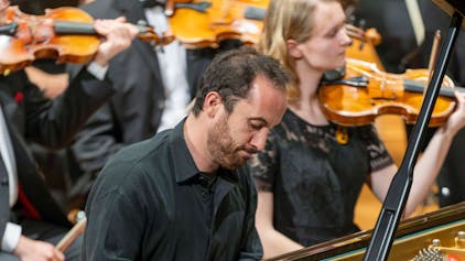 Auf dem Bild ist Igor Levit zu sehen, wie er beim Spielen konzentriert auf seinen Flügel schaut. Er trägt ein schwarzes Hemd. Im Hintergrund weitere Musikerinnen und Musiker.