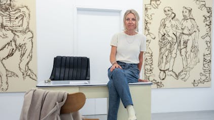 Die Galeristin Petra Martinetz sitzt auf einem Schreibtisch in ihrer Galerie, im Hintergrund sind Bilder zu sehen.