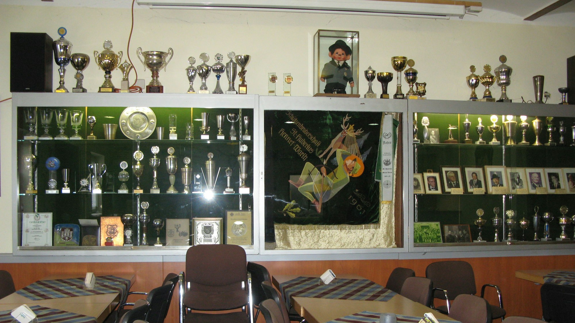 Das Bild zeigt die große Pokalvitrine der Hubertus-Schützen mit zahlreichen Trophäen, Standartentuch, Urkunden und Porträtfotos.