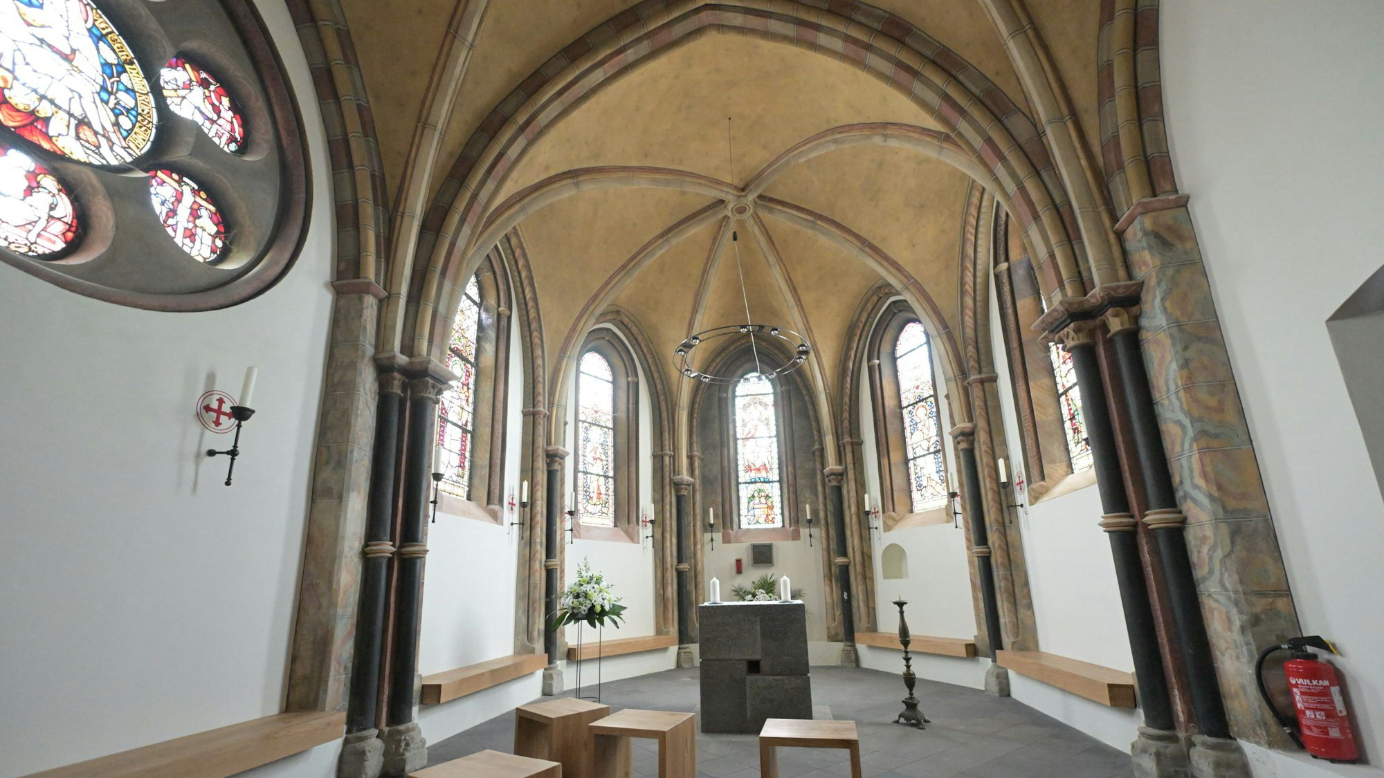 Innenraum der Markuskapelle Altenberg: Kreuzrippengewölbe, darunter fünf romanische Fenster, Sitzbänke und Hocker aus hellem Holt, davor ein Steinblock als Altar.