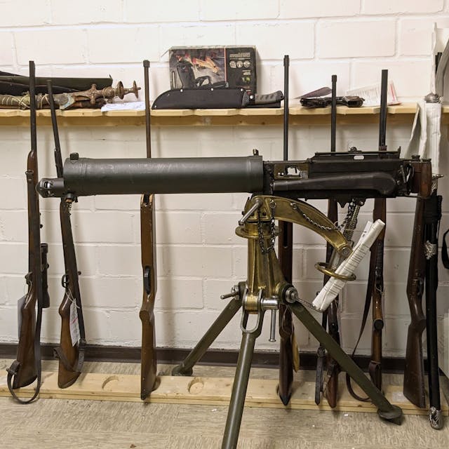 Ein altes Maschinengewehr ist auf einem Dreibein montiert. Dahinter lehnen diverse Waffen an der Wand.