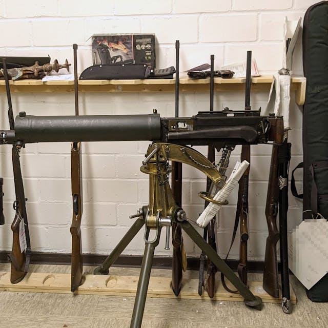 Ein altes Maschinengewehr ist auf einem Dreibein montiert. Dahinter lehnen diverse Waffen an der Wand.