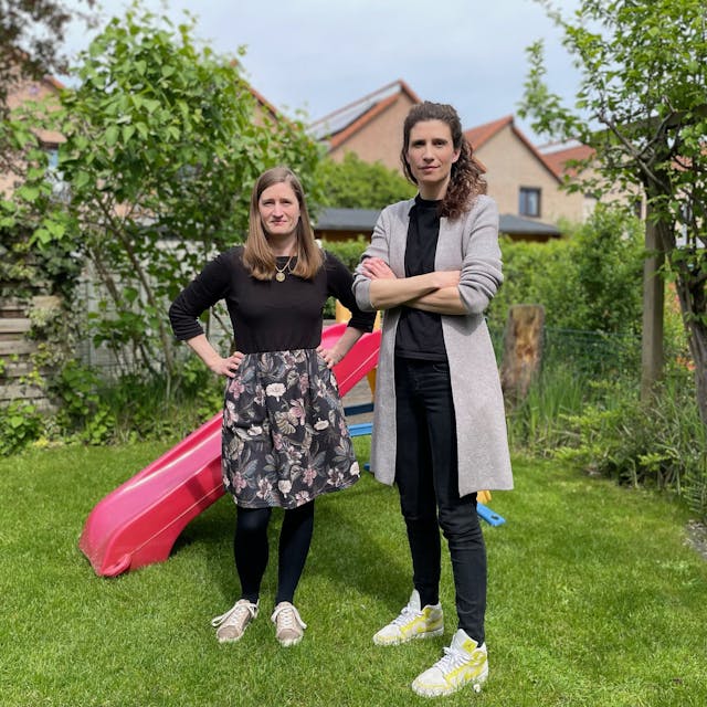 Zwei Frauen in einem Garten; im Hintergrund steht eine bunte Kinderrutsche aus Kunststoff.