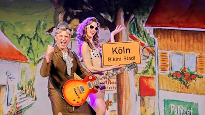 Schauspieler auf der Bühne: Eine Frau im Bikini und ein Mann als ältere Frau verkleidet halten ein Schild hoch auf dem steht: Köln, Bikini-Stadt.