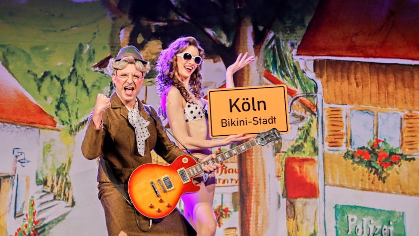 Schauspieler auf der Bühne: Eine Frau im Bikini und ein Mann als ältere Frau verkleidet halten ein Schild hoch auf dem steht: Köln, Bikini-Stadt.