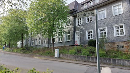 Ein mit grauem Schiefer verkleidetes Schulgebäude mit weißen Fensterrahmen. Vor dem Gebäude verläuft eine ansteigende Straße; auf dem Straßenschild steht Schulstraße