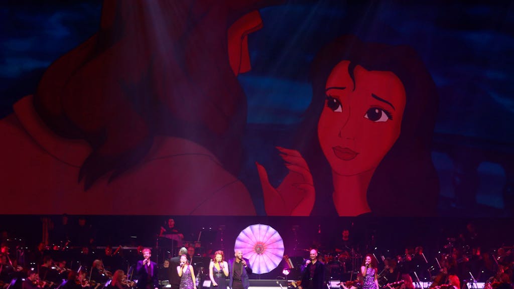 Ein Orchester mit Band und acht Sängern steht auf der Bühne. Im Hintergrund sieht man eine Szene aus Tarzan.&nbsp;