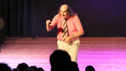 Guildo Horn steht auf der Bühne des Euskirchener Stadttheaters. Er trägt ein lachsfarbenes, kurzärmeliges Hemd mit einer braun-gestreiften Krawatte und eine helle Hose.