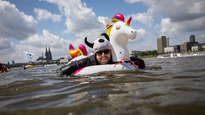 Ein kostümierter Teilnehmer schwimmt im Rhein am Panorama der Stadt mit dem Kölner Dom vorbei.&nbsp;
