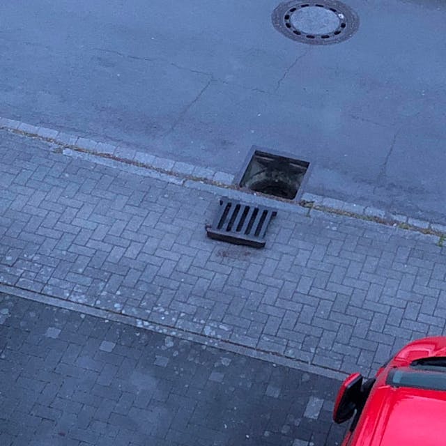 Auf dem Foto ist zu sehen, wie ein Gullydeckel nehmen dem Schacht auf dem Bürgersteig liegt.