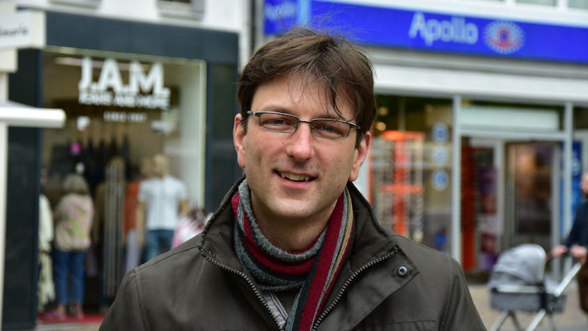 Ein Mann mit Brille, Schal und brauner Winterjacke vor Geschäften in einer Fußgängerzone.