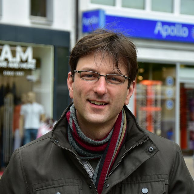 Ein Mann mit Brille, Schal und brauner Winterjacke vor Geschäften in einer Fußgängerzone.