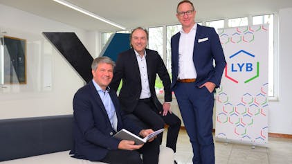 Drei Männer an und auf einem Sofa vor den Firmenlogos der beteiligten Unternehmen.