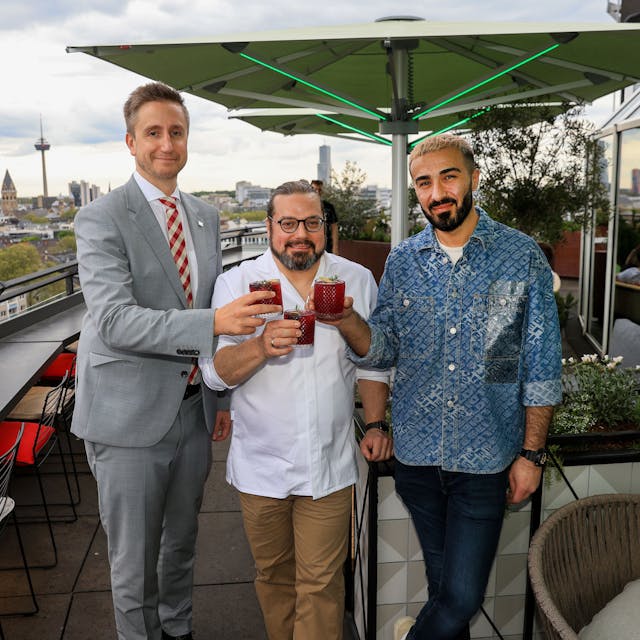 Drei Männer stehen mit Drinks in der Hand auf der Terrasse einer Rooftop-Bar.