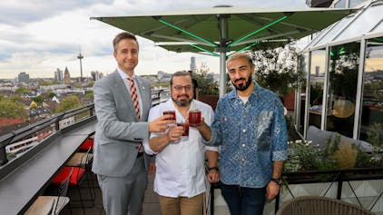 Drei Männer stehen mit Drinks in der Hand auf der Terrasse einer Rooftop-Bar.