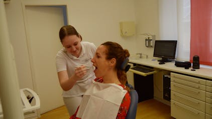 Celine Doerr blickt in der Zahnarztpraxis, in der sie arbeitet, in den Mund einer Patientin, die auf dem Behandlungsstuhl sitzt.
