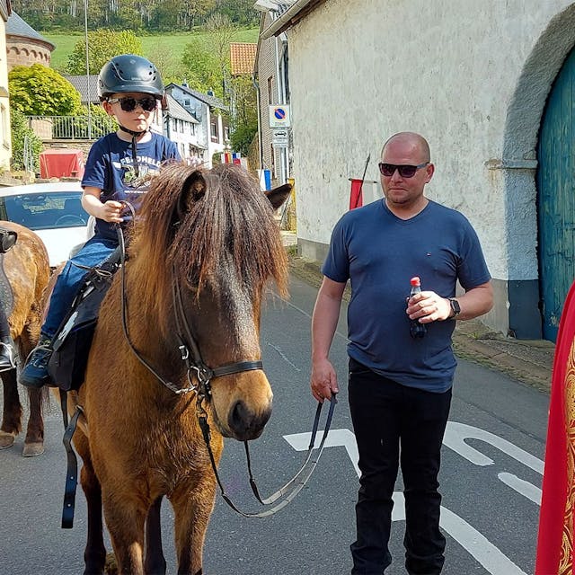 Zwei junge Teilnehmer reiten am Aachener Bischof in seinem roten Ornat vorbei. Eines der Pferde wird von einem Helfer geführt.