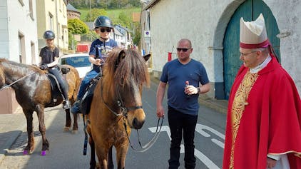 Zwei junge Teilnehmer reiten am Aachener Bischof in seinem roten Ornat vorbei. Eines der Pferde wird von einem Helfer geführt.