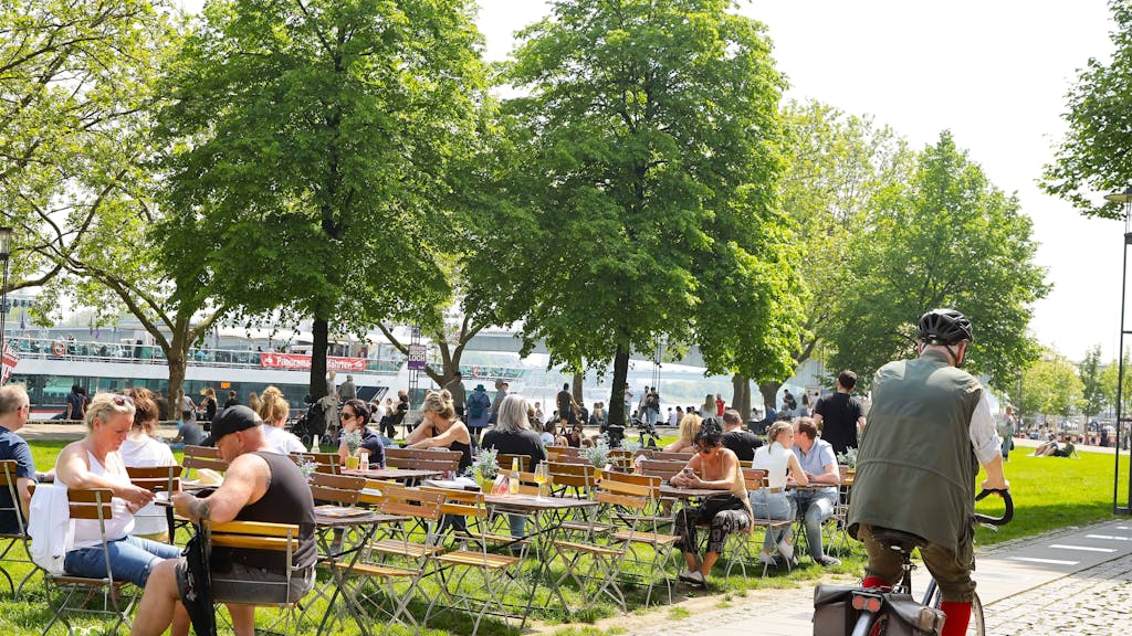 Menschen sitzen auf Gastro-Stühlen am Kölner Rheinufer in der Sonne.
