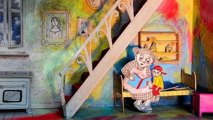 Eine Bilderbuchszene in einer Art kleiner Bühne. Eine aus Papier augeschnittene Frau sitzt mit einem kleinen Jungen auf dem Bett und liest ihm vor. Die Wände und Möbel sind in leuchtenden Farben gestaltet.