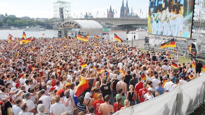 Eine Großleinwand steht auf der Deutzer Werft, im Hintergrund ist der Kölner Dom zu sehen. Auf der Fläche davor stehen viele hundert Fußballfans.