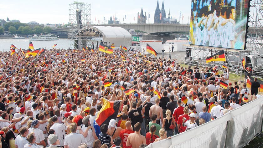 Eine Großleinwand steht auf der Deutzer Werft, im Hintergrund ist der Kölner Dom zu sehen. Auf der Fläche davor stehen viele hundert Fußballfans.